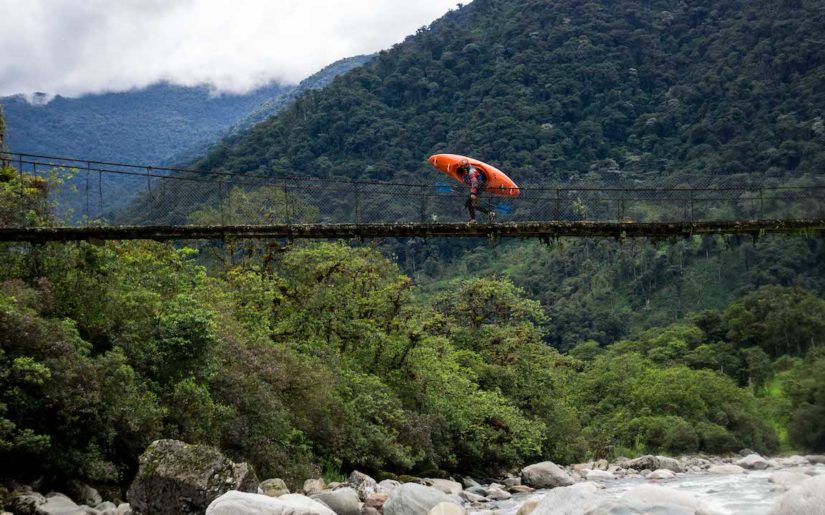 Man carrying kayak on suspended bridge, over an Ecuador river.